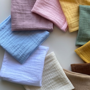 Top-down view of 11 folded organic muslin handkerchiefs arranged in a colorful circle on a pristine white background. Reusable double gauze cloth tissues in earthy and pastel minimalist tones.