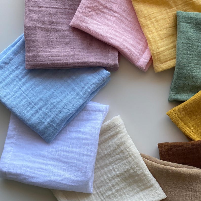 Top-down view of 11 folded organic muslin handkerchiefs arranged in a colorful circle on a pristine white background. Reusable double gauze cloth tissues in earthy and pastel minimalist tones.
