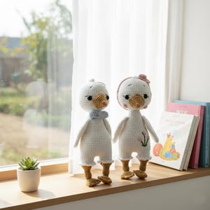 Amigurumi crochet goose dolls standing together on a sunlit window sill, with a vibrant green outdoor view and children's books. Perfect handmade decor for a nursery or child's bedroom.