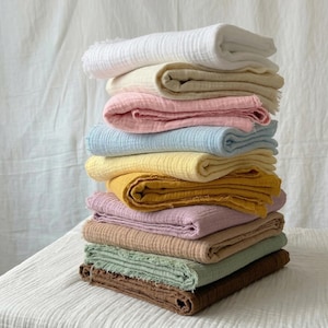 Puede incluir: Una pila de mantas de muselina dobladas en varios colores pastel, incluyendo blanco, crema, rosa, azul, amarillo y marrón. Las mantas tienen un aspecto texturizado y arrugado con bordes deshilachados. La pila está cuidadosamente organizada, mostrando los diferentes tonos.