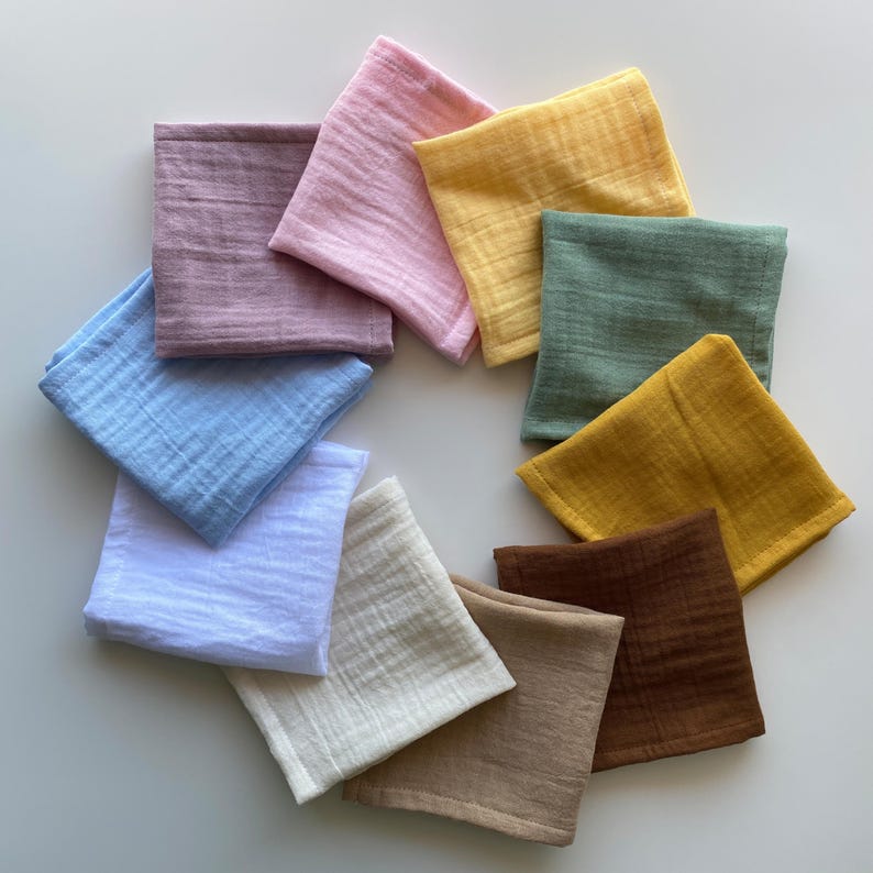 Circle arrangement of 11 organic muslin handkerchiefs in solid earthy and pastel colors. Reusable double gauze cotton hankies and zero waste unpaper tissues on a white background.