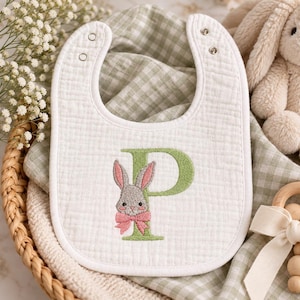 May include: A white muslin baby bib featuring an embroidered sage green letter P. A gray bunny with pink inner ears and a pink bow is stitched over the letter. The bib has a white trim and two silver-toned metal snap closures at the top neck area.