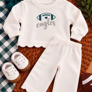 May include: A white long-sleeve sweatshirt and matching pants set with scalloped hems. The sweatshirt features a dark teal football graphic with a small heart and the word "eagles" embroidered in gray script. Includes white shoes with brown soles.