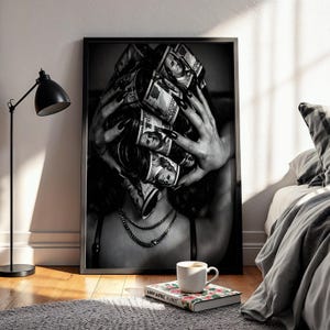 Peut inclure: Une impression d'art encadrée en noir et blanc représentant une personne les mains sur la tête, avec des rouleaux de billets de cent dollars américains en guise de bigoudis. La personne porte un collier à fine chaîne et un haut à bretelles noires.
