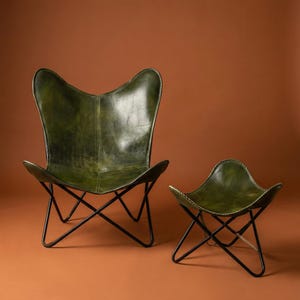Pode incluir: Uma cadeira borboleta de couro verde e um pufe a condizer. A cadeira e o pufe têm uma estrutura de metal preto. A cadeira tem uma forma distinta de asa de borboleta. O pufe é uma versão mais pequena da cadeira.