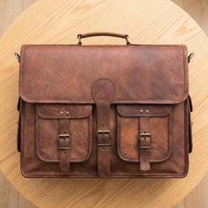 May include: A brown leather briefcase with two front pockets, each secured by a buckle. The bag has a top handle and a flap closure with a central strap and buckle. The leather has a distressed appearance.