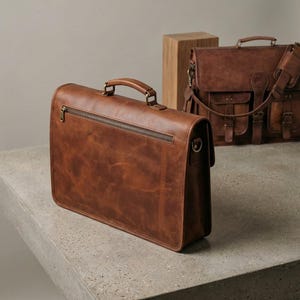 May include: A brown leather briefcase with a top handle and a zippered pocket on the back. The briefcase is rectangular and has a classic design. Another brown leather bag is in the background.