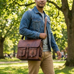 May include: A brown leather messenger bag with two front pockets and buckle closures. The bag has a flap closure and a shoulder strap. The bag is carried by a person wearing a denim jacket and khaki pants.