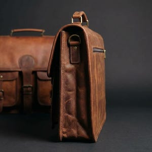 May include: A brown leather briefcase with a handle and shoulder strap attachments. The bag has a flap closure and a zipper pocket. Another leather bag is visible in the background.