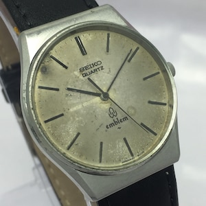 May include: A vintage Seiko Quartz watch with a silver-tone case and a black leather band. The watch face is a light gold color with black hour markers and hands. The word "emblem" is printed on the watch face.