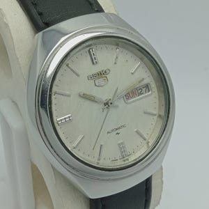 May include: Silver-toned Seiko 5 automatic watch with a white face, day and date display, and black leather strap. The watch face features silver hour markers and hands. The text "SEIKO 5 AUTOMATIC" is visible on the watch face.