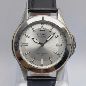 May include: Silver Casio Quartz watch with a black leather band. The watch face is silver with black hands and rectangular hour markers. The watch has the words "CASIO QUARTZ" and "WATER RESIST 50M" printed on the face.