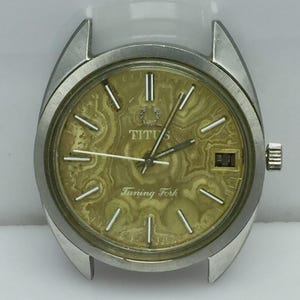 Op de afbeelding: Een vintage Titus horloge met een zilverkleurige kast en een getextureerde, geelgouden wijzerplaat. De wijzerplaat heeft zilveren uurmarkeringen, wijzers en de tekst "TITUS" en "Tuning Fork". Het horloge heeft een datumvenster op 3 uur.
