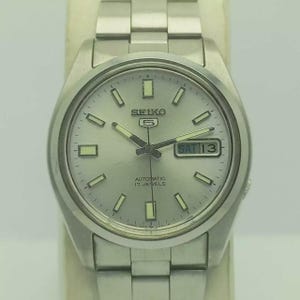 May include: A silver-toned Seiko 5 automatic wristwatch with a light gray dial. The watch features a day and date display, luminous hands and markers, and a stainless steel band. The watch face displays the text "SEIKO 5" and "AUTOMATIC 17 JEWELS".