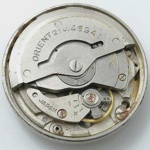 May include: A close-up of a watch movement with the word "ORIENT" and "JAPAN" visible. The movement is silver-colored with various gears, screws, and a balance wheel. The model number "21J.4694" is also visible.