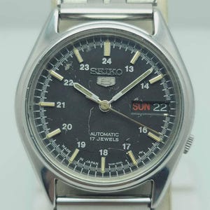 May include: A silver-toned Seiko automatic wristwatch with a black face. The watch features a day and date display, luminous hands and markers, and a stainless steel band. The watch face displays the text "SEIKO", "AUTOMATIC", "17 JEWELS", and "MADE IN JAPAN".