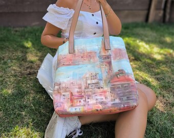 Könnte beinhalten: Eine große Tragetasche mit einem farbenfrohen Stadtlandschaftsgemälde. Die Tasche hat eine hellblaue und rosa Farbgebung mit einem hellbraunen Griff. Die Tasche wird von einer Person gehalten, die auf dem Gras sitzt.