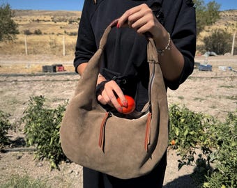 Könnte beinhalten: Olivgrüne Wildleder-Umhängetasche mit Reißverschluss und zwei braunen Lederquasten. Eine Person hält die Tasche, in der sich eine rote Tomate befindet. Der Hintergrund ist ländlich.