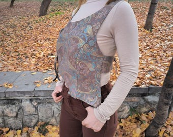 Könnte beinhalten: Eine braune Weste mit Paisley-Muster und blauen und goldenen Akzenten. Die Weste wird über einem beigefarbenen Rollkragenpullover getragen. Das Model trägt eine braune Hose. Der Hintergrund zeigt eine Steinmauer und herabgefallenes Herbstlaub.