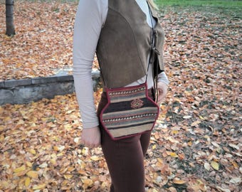 Könnte beinhalten: Eine braune Wildlederweste und eine kleine, gemusterte Umhängetasche mit rotem Rand. Die Tasche hat ein geometrisches Design mit Streifen in Braun-, Beige- und Grautönen. Der Hintergrund zeigt fallende Blätter.
