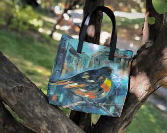 Könnte beinhalten: Eine Tragetasche mit einem Gemälde eines Vogels auf einem Ast. Der Vogel hat schwarze, orange und gelbe Federn. Der Hintergrund ist blau und türkis mit einem Gebäudedesign. Die Tasche hat schwarze Griffe.
