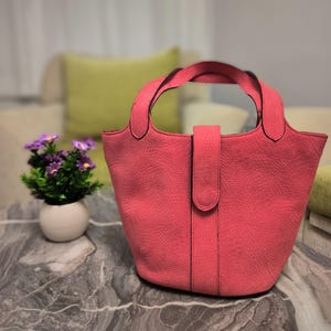 Könnte beinhalten: Eine korallenfarbene Handtasche mit einem strukturierten, reptilienartigen Muster. Die Tasche hat zwei Griffe und einen zentralen Riemenverschluss. Die Tasche steht auf einer Marmoroberfläche, mit einer kleinen Topfpflanze mit lila Blumen im Hintergrund.
