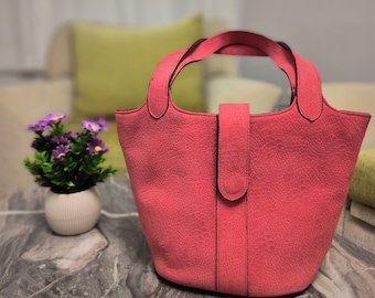 Könnte beinhalten: Eine korallenfarbene Handtasche mit einem strukturierten, reptilienartigen Muster. Die Tasche hat zwei Griffe und einen zentralen Riemenverschluss. Die Tasche steht auf einer Marmoroberfläche, mit einer kleinen Topfpflanze mit lila Blumen im Hintergrund.