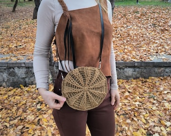 Könnte beinhalten: Eine runde, gewebte, braune Handtasche mit radialem Muster. Die Tasche hat einen schwarzen Riemen und wird quer über dem Körper getragen. Der Hintergrund zeigt abgefallene Herbstblätter.
