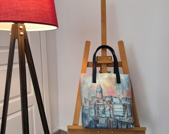 Könnte beinhalten: Eine Tragetasche mit einem farbenfrohen Stadtbilddesign, das einen Turm und Gebäude in Blau-, Orange- und Rosatönen zeigt. Die Tasche hat schwarze Griffe und wird auf einer hölzernen Staffelei ausgestellt. Ein roter Lampenschirm ist im Hintergrund zu sehen.