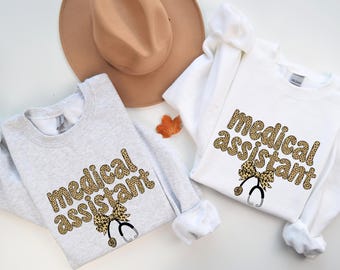 May include: Two long-sleeved sweatshirts, one light gray and one white, each with the words "medical assistant" in a leopard print font. A stethoscope and bow graphic is below the text. A tan hat and fall leaf are in the background.