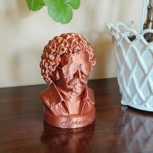 May include: A metallic copper-coloured bust of a person with curly hair, a moustache, and sunglasses, wearing a collared shirt. The base of the sculpture features the embossed text Moroder in a cursive script, capturing the likeness of the individual.