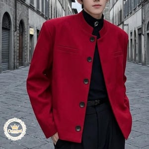 Könnte beinhalten: Eine leuchtend rote Jacke mit Stehkragen und vier dunklen Knöpfen. Die Jacke hat zwei kleine Taschen und wird über einem schwarzen Rollkragenpullover und einer schwarzen Hose getragen. Die Jacke ist einfarbig und hat eine taillierte Passform.