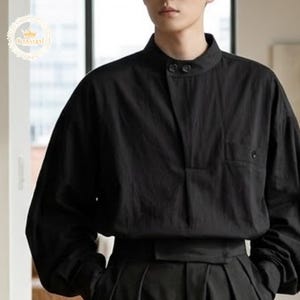 Può includere: Camicia nera a maniche lunghe con colletto alla coreana e due bottoni. La camicia ha una vestibilità ampia e una piccola tasca sul petto. La camicia è realizzata in tessuto leggero.