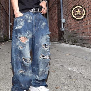 May include: Distressed blue denim jeans with multiple rips and tears. The jeans have a loose fit and are worn with a black belt and white trainers. The image is set against a brick wall background. The jeans are approximately 100 cm in length.