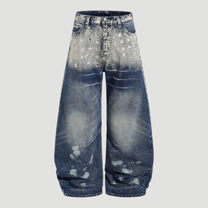 May include: Blue denim jeans with a wide leg design. The jeans feature a faded wash with distressed details, including circular cutouts near the waist and white paint splatters. The jeans have a classic five-pocket design.