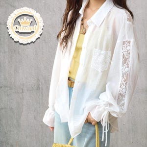 Op de afbeelding: Witte blouse met kanten details op de mouwen en de zak. De blouse heeft een kraag en lange mouwen met strikmanchetten. Het wordt gedragen over een gele top en lichtblauwe jeans. De afbeelding bevat ook een decoratief bord met het woord "Crowned".