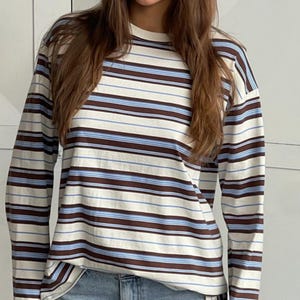 Puede incluir: Una camiseta de manga larga con cuello redondo que presenta un patr&oacute;n de rayas horizontales en tonos marr&oacute;n, azul claro y crema. La prenda tiene un corte holgado con mangas largas y un cuello acanalado, mostrando un dise&ntilde;o de rayas constante.
