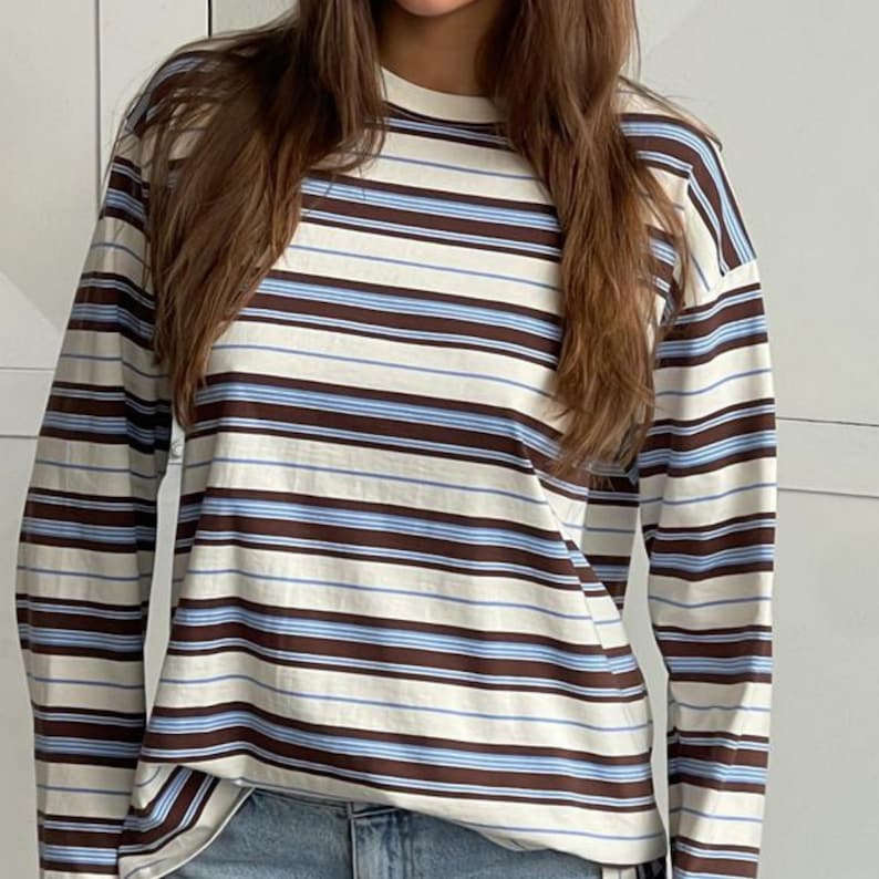 Puede incluir: Una camiseta de manga larga con cuello redondo que presenta un patr&oacute;n de rayas horizontales en tonos marr&oacute;n, azul claro y crema. La prenda tiene un corte holgado con mangas largas y un cuello acanalado, mostrando un dise&ntilde;o de rayas constante.