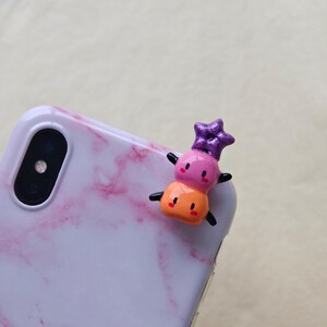 K&ouml;nnte beinhalten: Ein farbenfroher Telefonanh&auml;nger mit einem gestapelten Design und einem Stern oben. Der Anh&auml;nger hat ein rosa, orange und lila Farbschema mit schwarzen Akzenten. Der Anh&auml;nger ist an einer wei&szlig;en Handyh&uuml;lle mit rosa Marmormuster befestigt.
