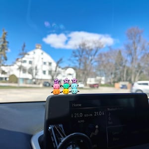 K&ouml;nnte beinhalten: Vier bunte, gestapelte schneemannartige Figuren mit sternf&ouml;rmigen H&uuml;ten sitzen auf dem Armaturenbrett eines Autos. Die Figuren sind rosa, orange, gr&uuml;n und blau. Die Anzeige des Autos zeigt 21,0 Grad Celsius an.
