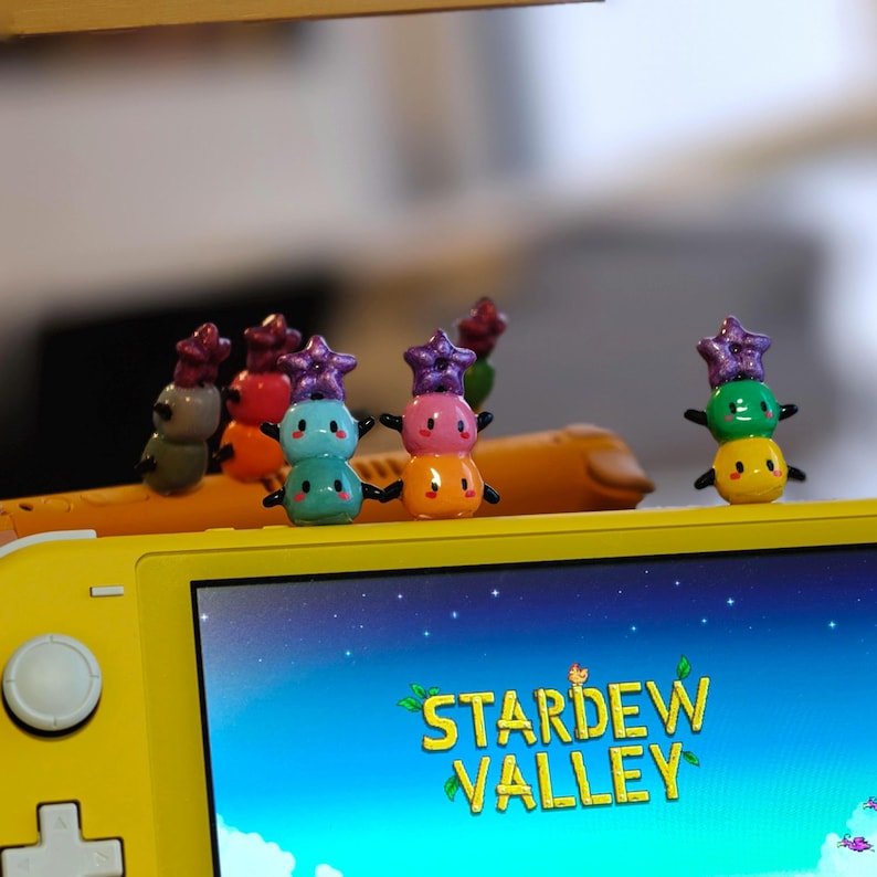 K&ouml;nnte beinhalten: Eine Sammlung bunter, gestapelter Schneemannfiguren mit sternf&ouml;rmigen H&uuml;ten, die auf einer gelben Spielekonsole liegen. Auf dem Bildschirm ist das Logo des Spiels "Stardew Valley" zu sehen. Die Figuren sind in verschiedenen Farben gehalten, darunter Gr&uuml;n, Rosa und Blau.