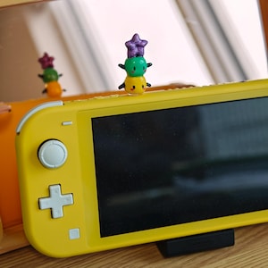 K&ouml;nnte beinhalten: Eine gelbe Nintendo Switch Lite-Konsole mit einer kleinen, bunten Figur oben drauf. Die Figur ist ein Stapel aus drei runden Formen: gelb, gr&uuml;n und lila, mit kleinen schwarzen Armen. Die Konsole steht auf einem schwarzen St&auml;nder.