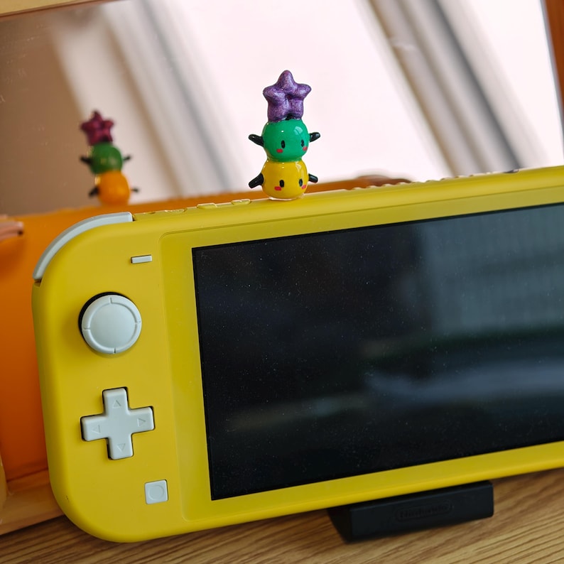 K&ouml;nnte beinhalten: Eine gelbe Nintendo Switch Lite-Konsole mit einer kleinen, bunten Figur oben drauf. Die Figur ist ein Stapel aus drei runden Formen: gelb, gr&uuml;n und lila, mit kleinen schwarzen Armen. Die Konsole steht auf einem schwarzen St&auml;nder.