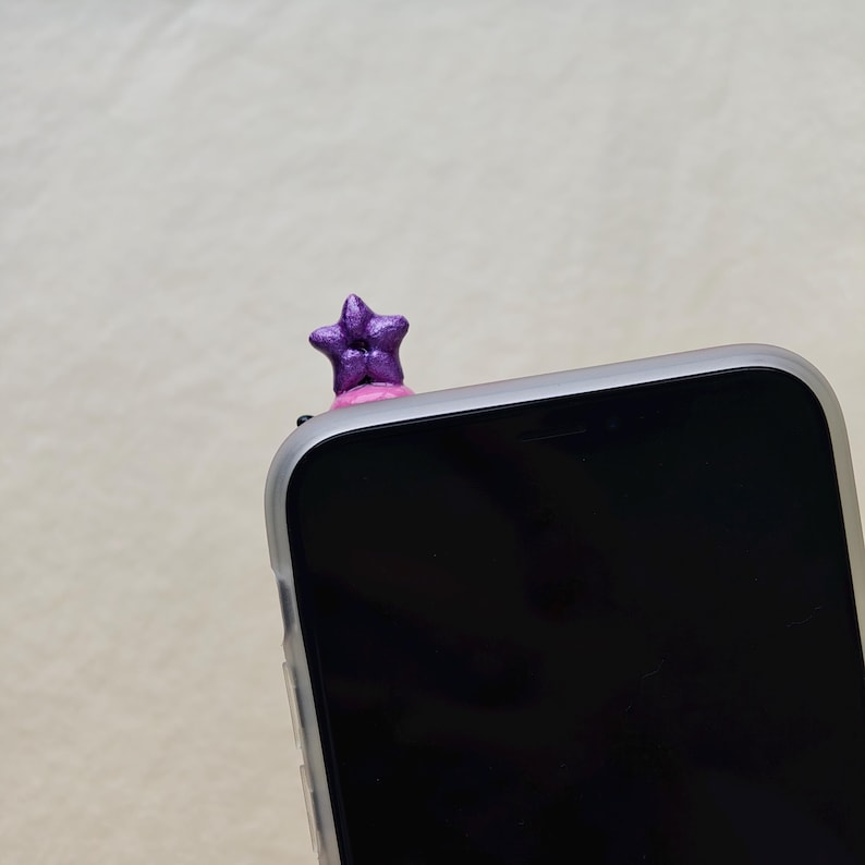 K&ouml;nnte beinhalten: Ein lila sternf&ouml;rmiger Telefonanh&auml;nger sitzt auf einem Smartphone. Der Stern hat eine strukturierte Oberfl&auml;che und ist auf einer rosa Basis montiert. Das Telefon befindet sich in einer transparenten H&uuml;lle.