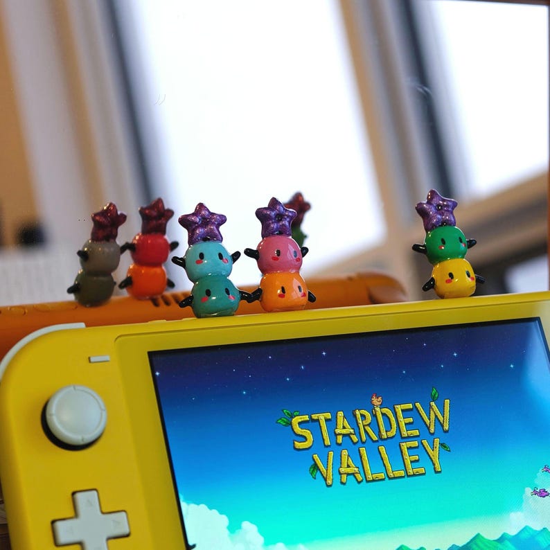 K&ouml;nnte beinhalten: Eine gelbe Nintendo Switch Lite-Konsole, die das Spiel "Stardew Valley" anzeigt. Mehrere bunte, gestapelte Figuren mit sternf&ouml;rmigen H&uuml;ten sind oben auf der Konsole angeordnet und verleihen dem Gaming-Setup eine spielerische Note.