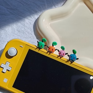 May include: Four colorful, cartoon-like figures with green leaf tops are arranged on a yellow handheld gaming device. The figures are in green, orange, pink, and blue. The device is on a white surface with a curved edge.