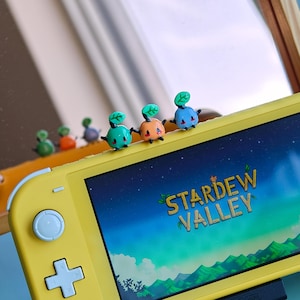 May include: A yellow handheld gaming console displaying the Stardew Valley title screen. Perched on the top edge are six small, colourful, glossy figurines shaped like round creatures with tiny leaves on their heads in green, orange, and blue.