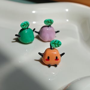 May include: Three small, glossy, fruit-shaped figurines with tiny black limbs and a single green leaf on top. One figurine is bright green, one is light purple, and the front-most one is orange with two small red dots on its face.