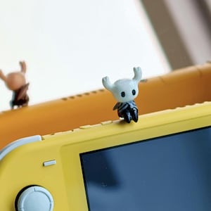May include: A small, white and black figurine of a character with horns, placed on a yellow handheld gaming device. Another similar figurine is visible in the background. The figurine has a detailed design.