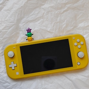K&ouml;nnte beinhalten: Eine leuchtend gelbe Handheld-Spielkonsole mit schwarzem Bildschirm. Eine kleine, bunte Figur aus gestapelten Charakteren sitzt obenauf. Die Konsole verf&uuml;gt &uuml;ber ein Steuerkreuz, Tasten und Steuerkn&uuml;ppel. Der Hintergrund ist eine strukturierte wei&szlig;e Oberfl&auml;che.